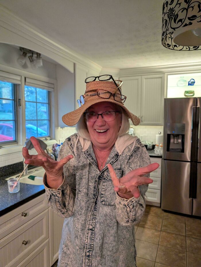 Older woman in kitchen wearing multiple glasses and a hat, smiling happily in wholesome mom moments.