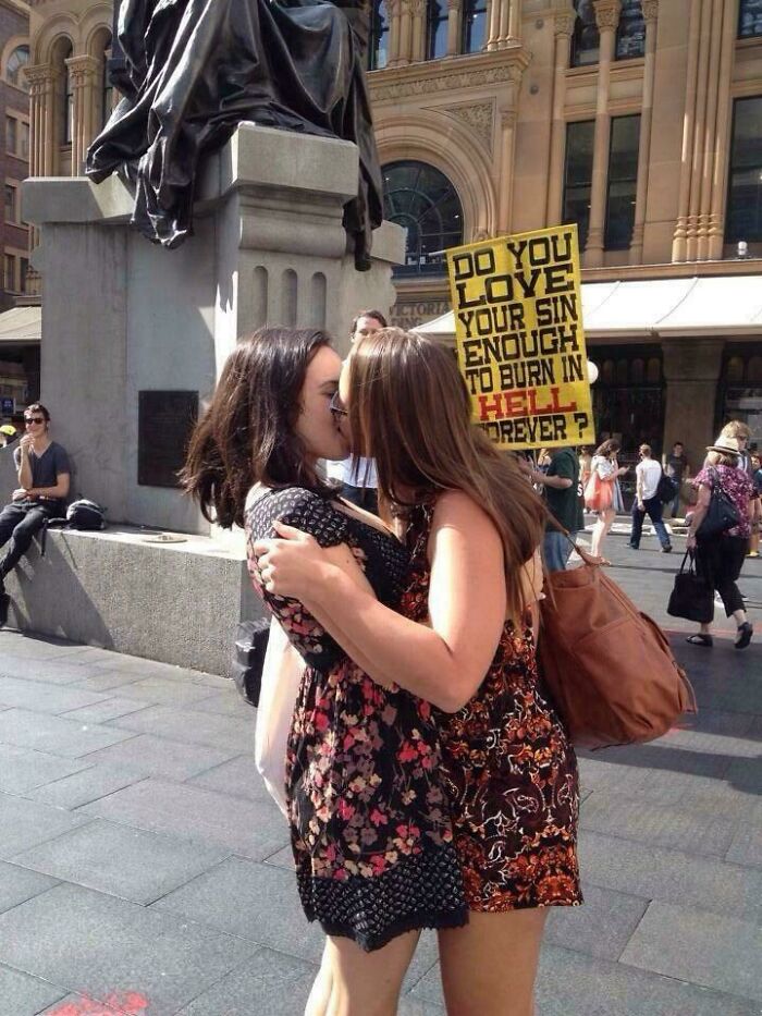 Two women kissing in public as a peaceful act of chaotic good against a harsh protest sign about sin and hell.