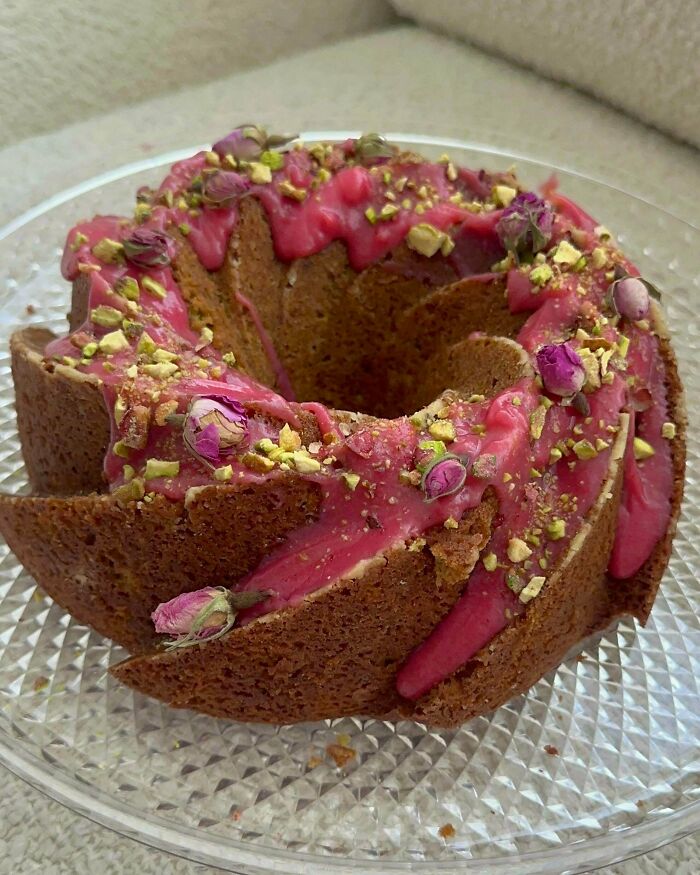 Bundt cake with pink glaze, pistachios, and dried rose buds on a clear glass plate showcasing gorgeous baked goods.