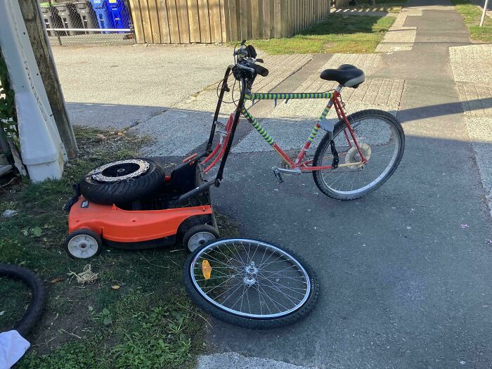 A redneck engineer's inventive lawnmower bike hybrid with a detached front bicycle wheel on a driveway.