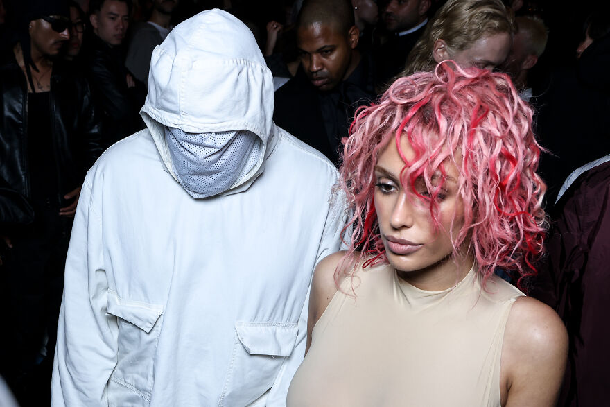 Bianca Censori with pink hair and West wearing a white hooded jacket at an event in a dark crowded setting. Bianca Censori with pink hair and West wearing a white hooded jacket at an event in a dark crowded setting.
