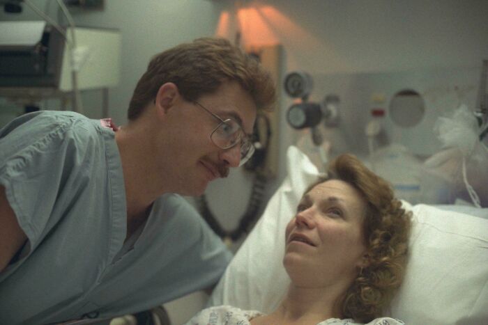 Man in hospital gown smiling at woman lying in hospital bed, capturing a wholesome moment parents and kids share.