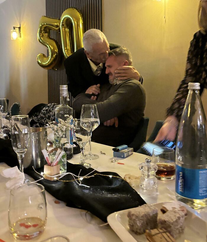 Elderly man affectionately hugging younger man at a dinner table, capturing funny and wholesome parent moments.