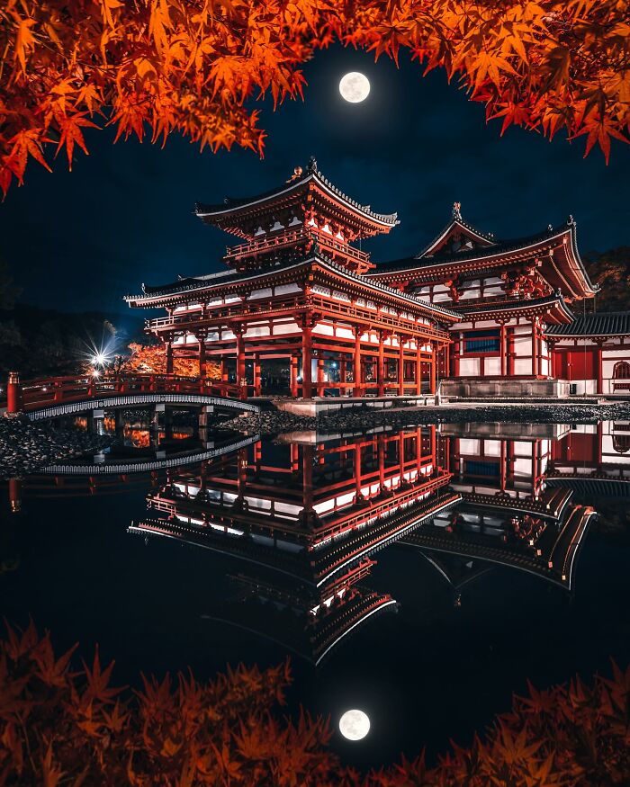 Traditional Asian temple illuminated at night with full moon and autumn leaves, reflecting in calm water.