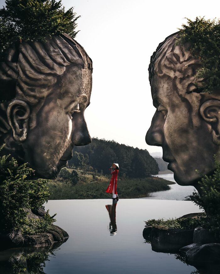 Large stone faces with greenery and a person in red standing by water, capturing glimpses of everyday life in Asia.