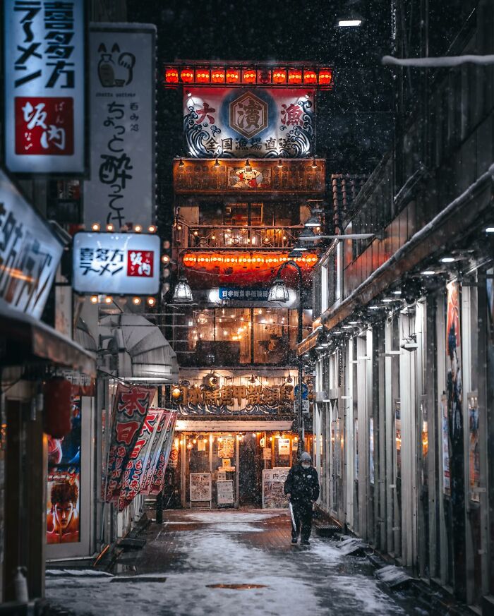 Snowy night street scene in Asia with illuminated signs and a lone pedestrian walking through the quiet alleyway.