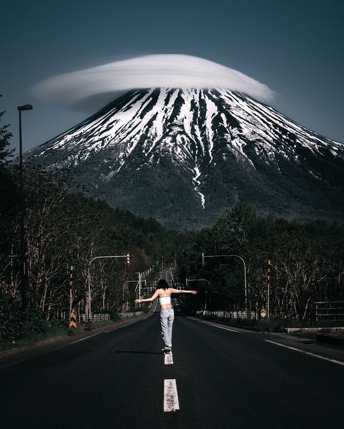 Person skateboarding down an empty road with a snow-capped mountain and cloud formation in everyday life in Asia.
