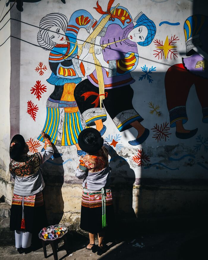 Two people in traditional attire painting a colorful mural depicting Asian cultural scenes, everyday life in Asia.
