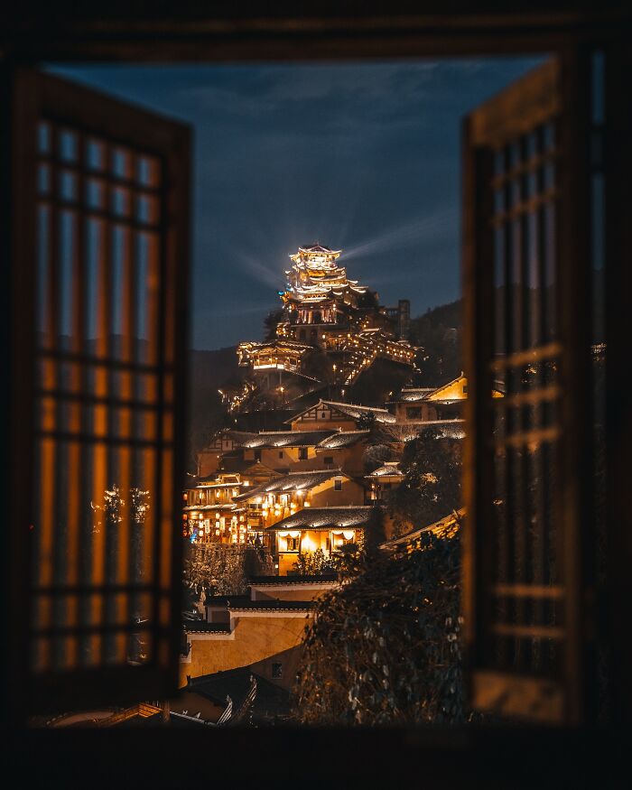 Night view of illuminated Asian hillside village and temples framed by traditional window, capturing glimpses of everyday life in Asia.
