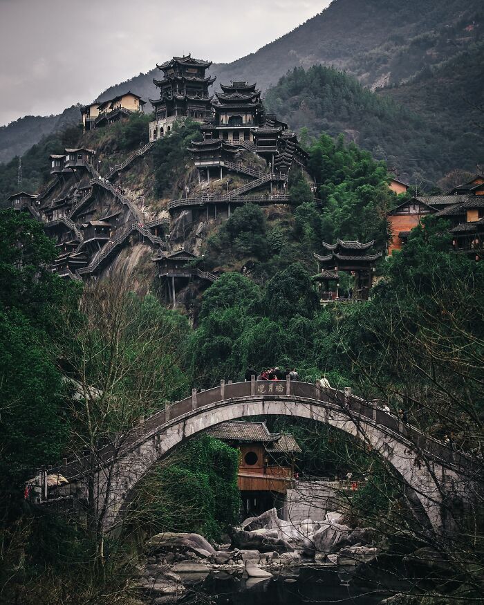 Ancient Asian architecture on a mountainside surrounded by lush greenery and a stone bridge over a river in everyday life.