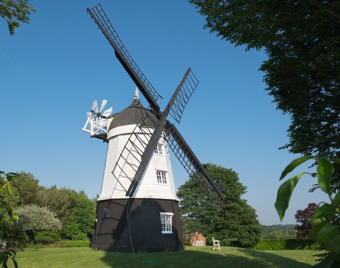 Historic windmill house with black and white exterior surrounded by greenery under clear blue sky, iconic TV houses in real life