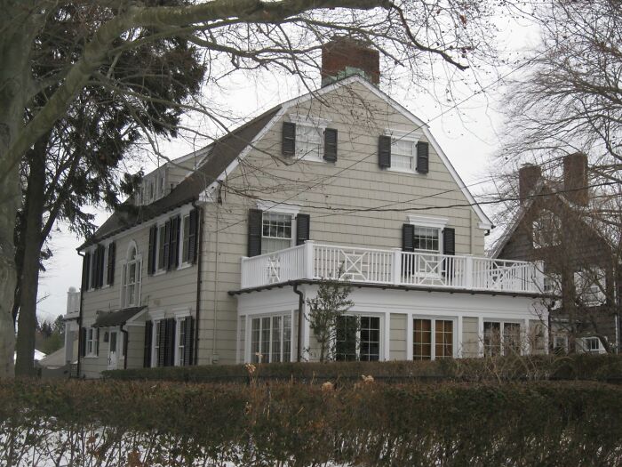Large iconic TV house with white siding and black shutters surrounded by leafless trees and winter shrubs.