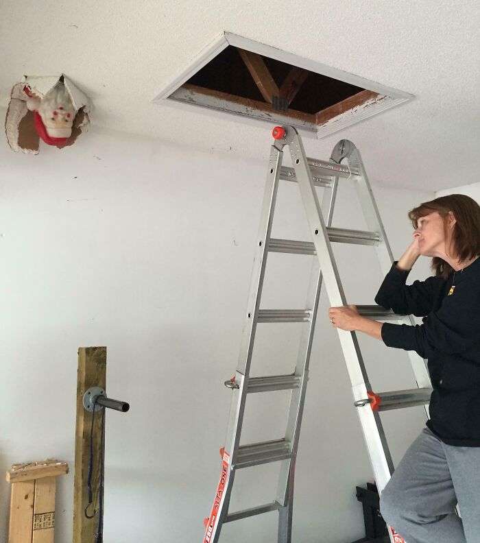Woman looking thoughtfully at a ladder under an attic opening with a Santa decoration stuck in the ceiling hole, funny wholesome parents moment.