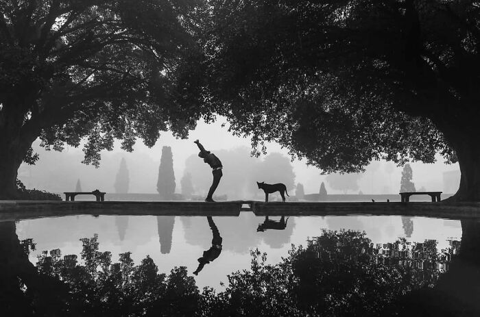 Black and white photo capturing everyday life in India with a person stretching near a dog by a reflective water body.
