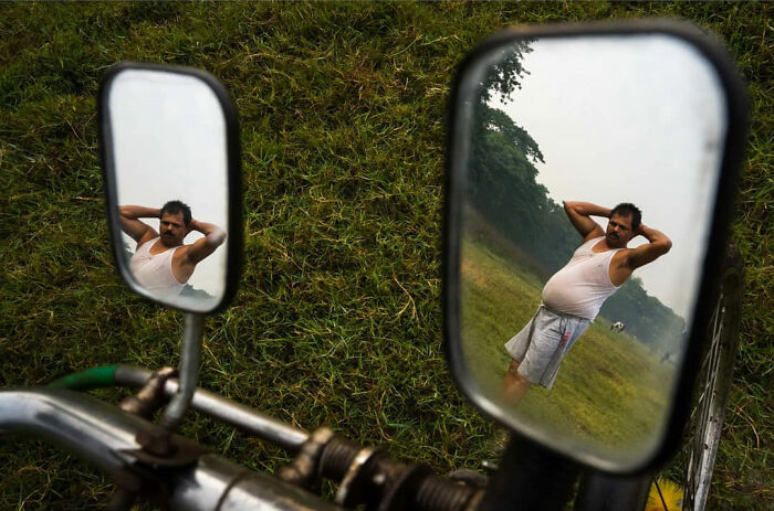 Man in white tank top and shorts relaxing in grassy field, reflected in motorcycle mirrors, capturing everyday life in India.