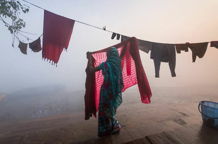 Woman in traditional attire hanging clothes on a line in foggy morning, capturing everyday life in India.