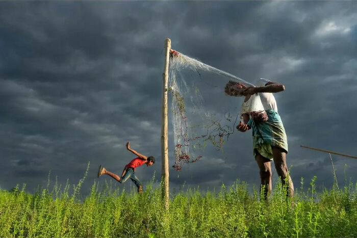 Man mending fishing net and child playing in green field under dramatic cloudy sky, capturing everyday life in India.
