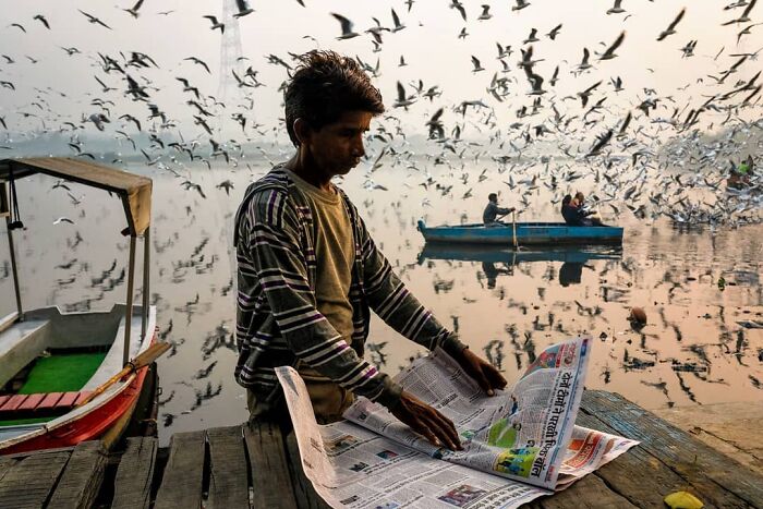 Young man reading a newspaper by the riverbank with birds flying overhead, capturing everyday life in India.