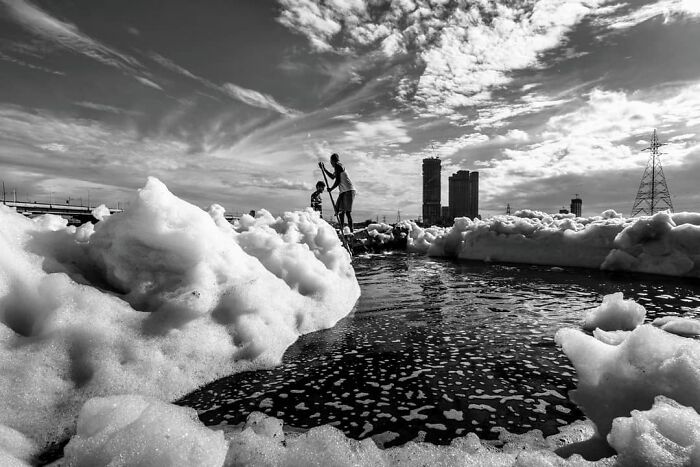 Two people standing among thick foam near water, depicting everyday life in India with city buildings in background.