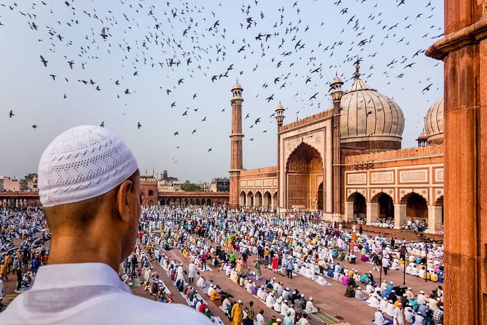 Man wearing white cap overlooking a large crowd gathered for prayer at a historic mosque, capturing everyday life in India.