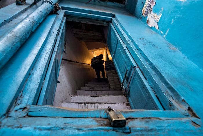 Man carrying boxes down dimly lit stairs inside a blue-painted building, capturing everyday life in India.