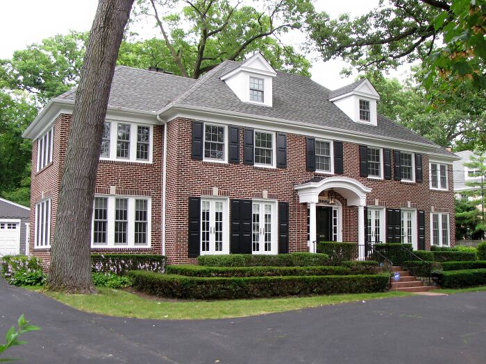 Large brick TV house with white-trimmed windows and black shutters surrounded by trees and greenery in real life setting