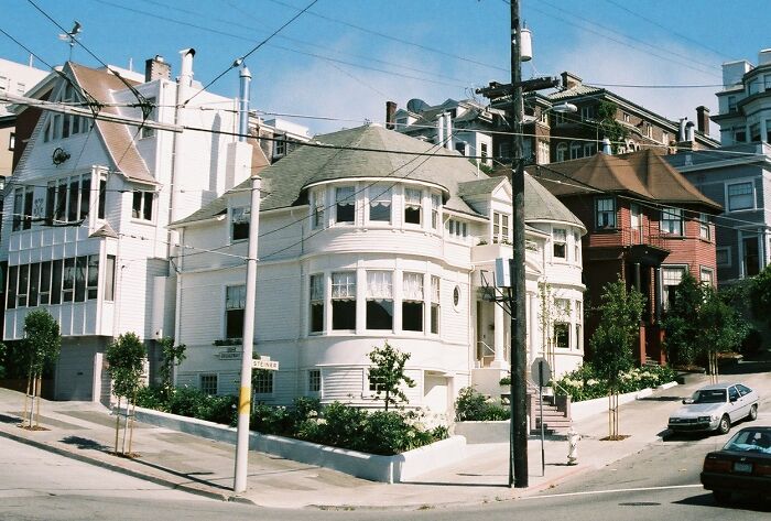 Victorian-style iconic TV house situated on a sloped street with parked cars and clear blue sky in the background.