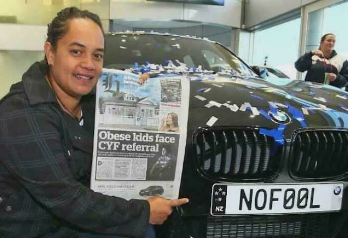 Person pointing to a newspaper headline while sitting next to a black car with a custom license plate in an indoor setting.