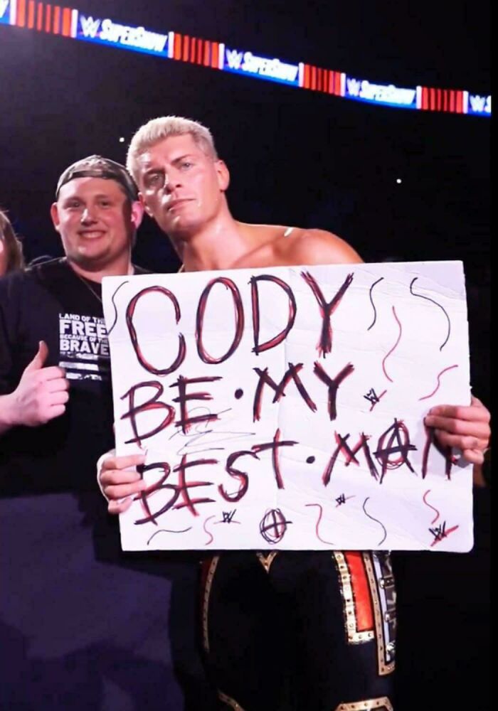 Two men at a wrestling event, one holding a handmade sign, inspiring facts better than watching the news.