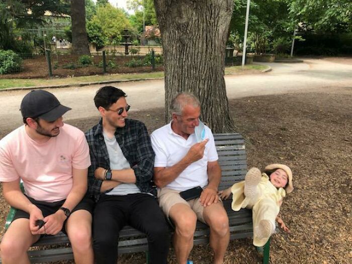 Three men sitting on a bench in a park while a child slips off the edge, capturing a perfect moment before disaster struck.