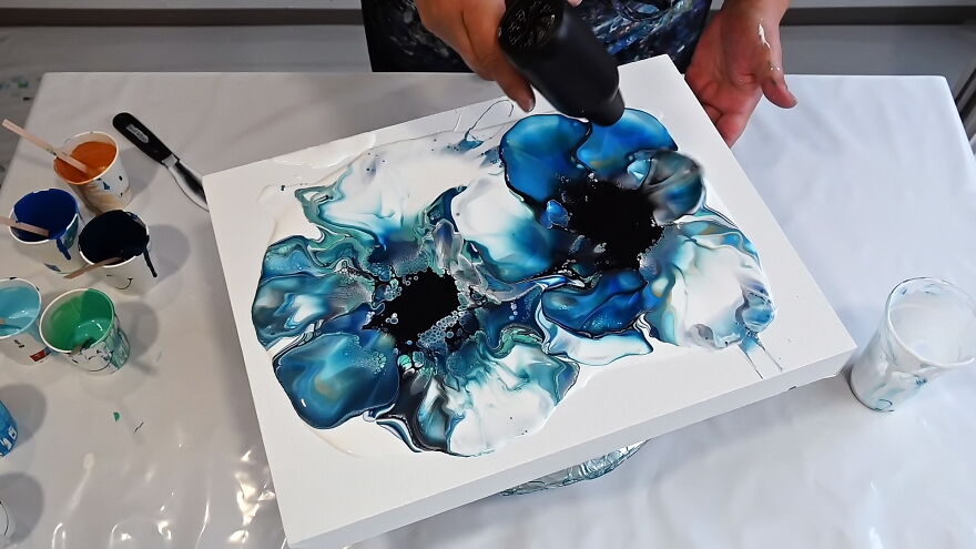 Acrylic pouring creating a deconstructed bloom with blue and gold colors on a white canvas using a heat tool.