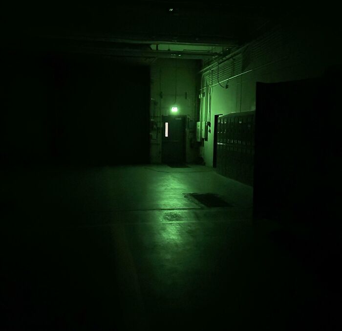 Dimly lit empty industrial hallway with green emergency exit light, illustrating creepy and unsettling jobs that should pay more.