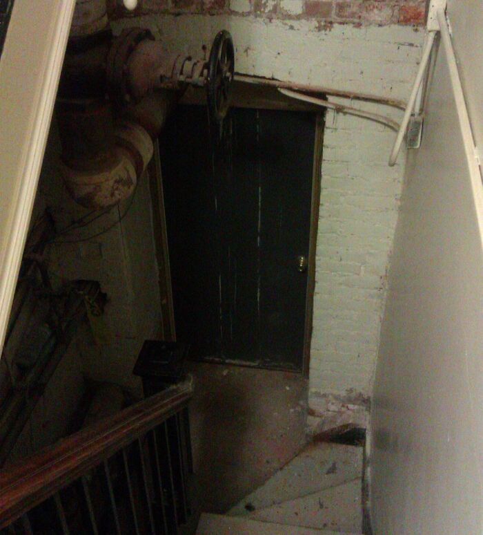 Dimly lit basement staircase leading to a dark, old door and rusty pipes in a creepy work environment photo.