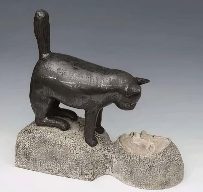 Bronze medieval cat sculpture standing on a c*****d stone human face, illustrating historical human fascination with cats.