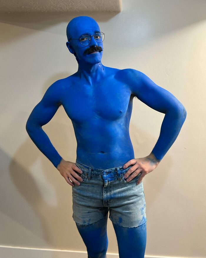 Man in genius Halloween costume with blue body paint, mustache, glasses, and denim shorts posing confidently indoors.