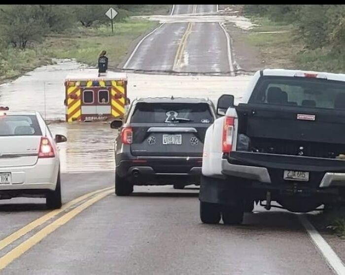 Ambulance partially submerged in floodwater while cars and trucks wait on the road, related to EMT and paramedic humor.