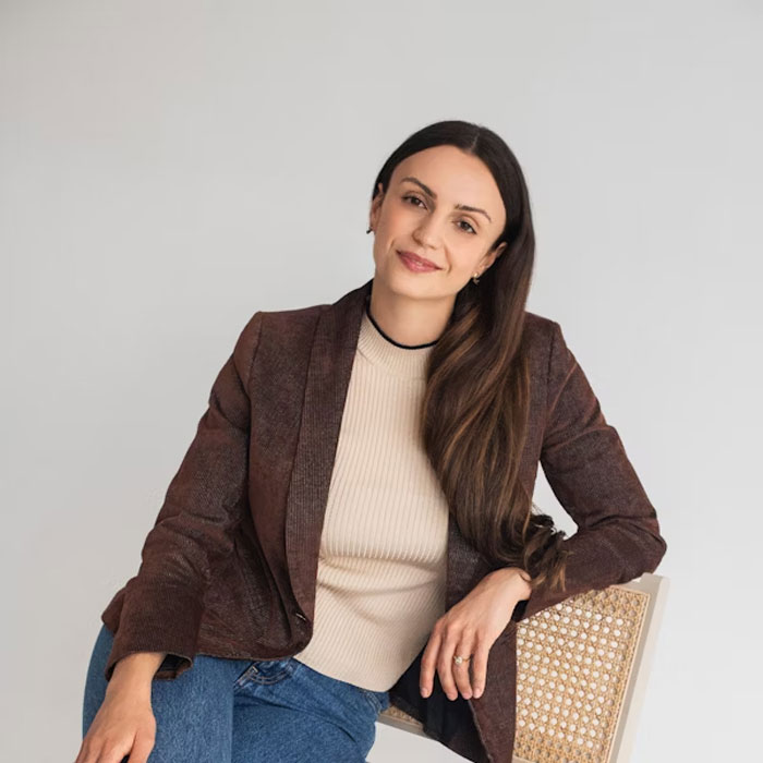Young woman with long dark hair wearing a brown blazer and beige top, seated on a chair in a relaxed pose. Young woman with long dark hair wearing a brown blazer and beige top, seated on a chair in a relaxed pose.