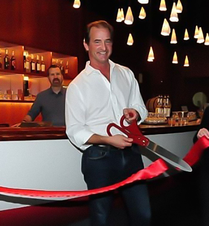 Man holding large scissors cutting red ribbon at event, related to husband of woman caught with CEO amid divorce news. Man holding large scissors cutting red ribbon at event, related to husband of woman caught with CEO amid divorce news.