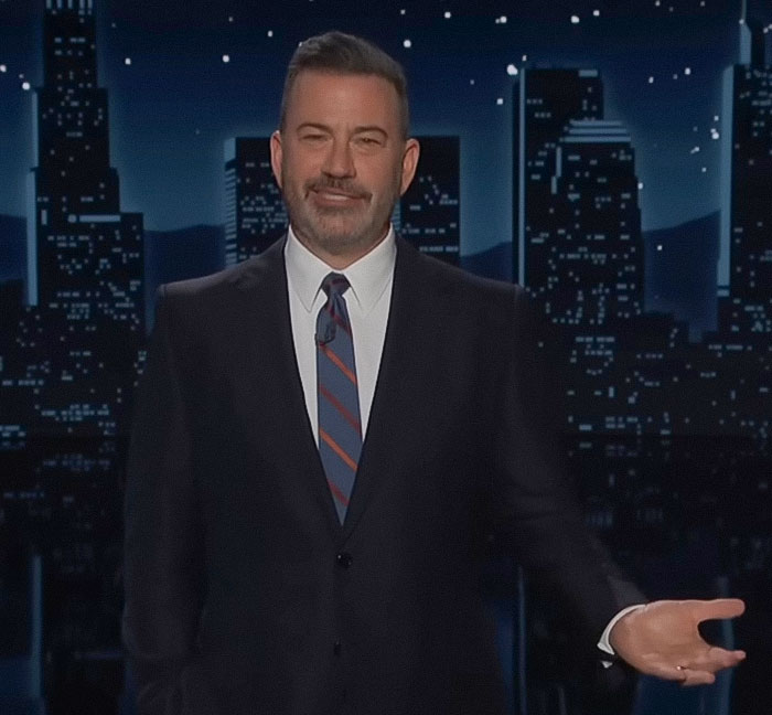 Jimmy Fallon speaking on stage in a suit and tie against a nighttime city skyline background during a show. Jimmy Fallon speaking on stage in a suit and tie against a nighttime city skyline background during a show.