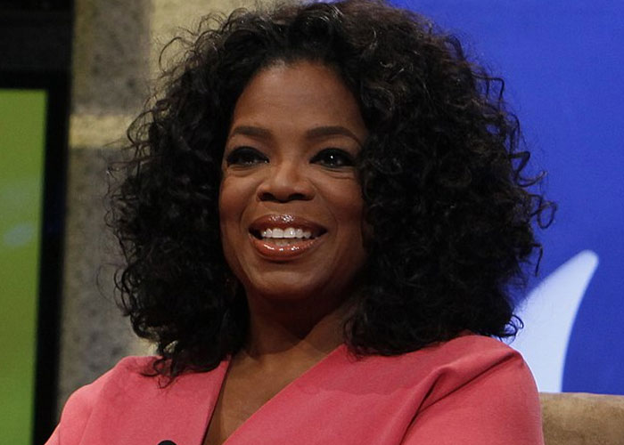 A woman with curly hair and a pink blazer smiling while discussing historical events with societal damage impact.