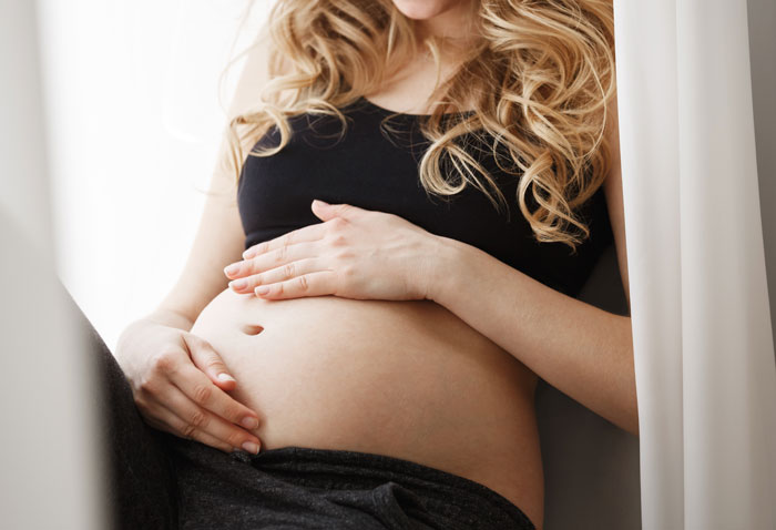 Pregnant woman gently touching her belly near a window, capturing a quiet and reflective moment at home.