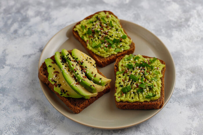 Three slices of toasted bread topped with mashed and sliced avocado seasoned with seeds and herbs on a plate.
