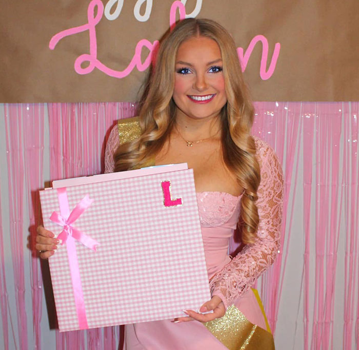 Cheerleader in pink dress smiling and holding a large pink gift box with ribbons at an event background. Cheerleader in pink dress smiling and holding a large pink gift box with ribbons at an event background.