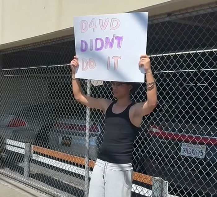 Young man holding a sign near an abandoned Tesla linked to singer, amid decomposing body discovery controversy. Young man holding a sign near an abandoned Tesla linked to singer, amid decomposing body discovery controversy.