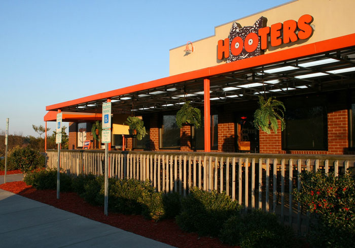Hooters restaurant exterior with orange sign, wooden fence, and hanging plants on a clear sunny day.