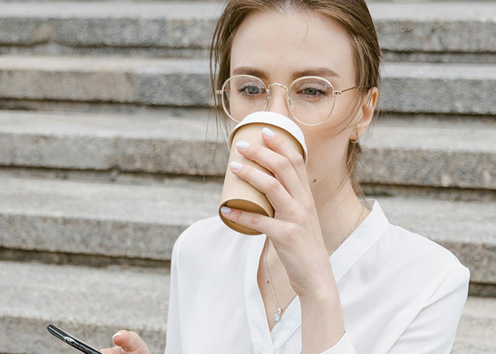 Young woman with glasses sipping coffee outdoors while checking phone, illustrating common purchases ditched due to ridiculous price tags.