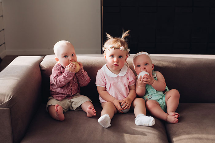 Three babies sitting on a couch, two holding round toys, siblings showing family ties and contrast in lifestyles.