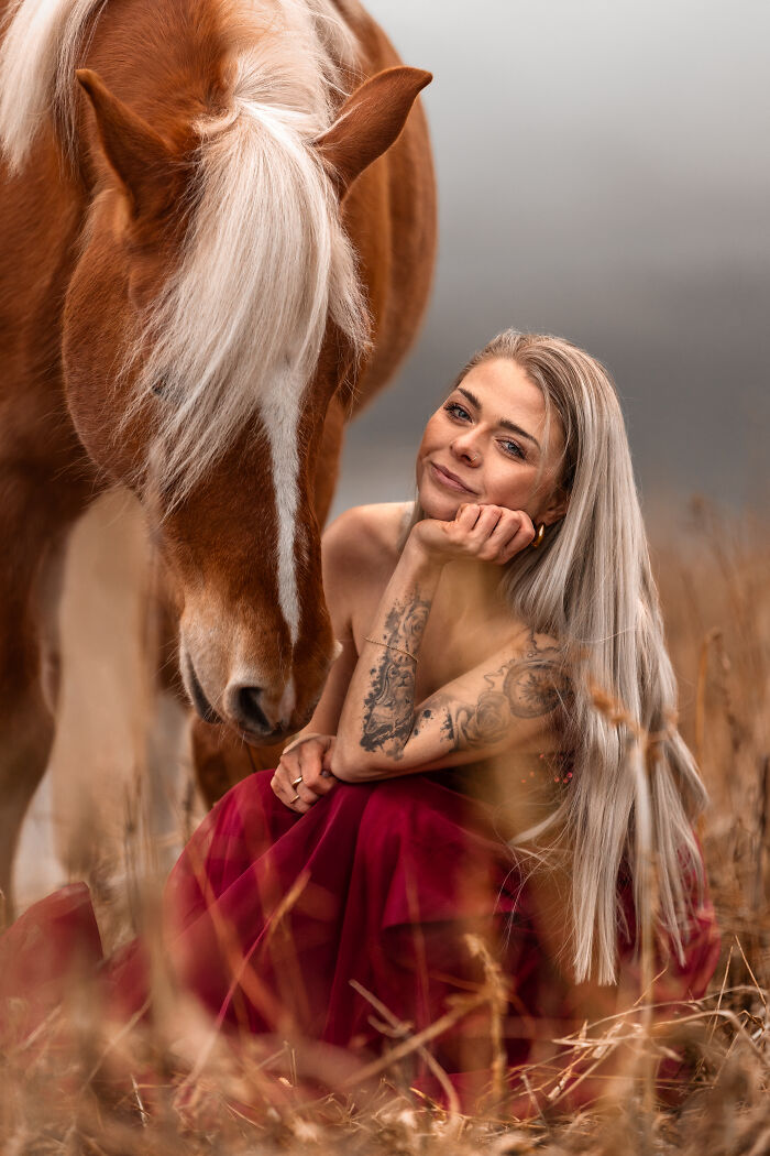 Woman with long blonde hair and tattoos sitting in a field beside a horse in a creative pet photo winning award.