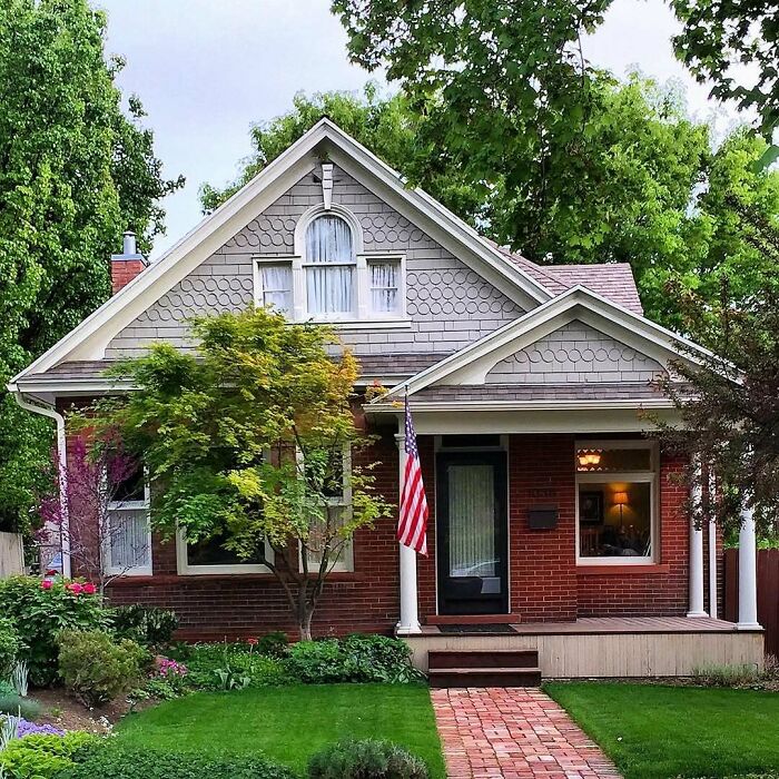 Charming old home with brick exterior, detailed shingles, lush greenery, and an inviting front porch with an American flag.