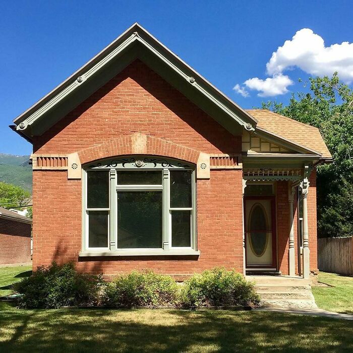 Brick old home with decorative trim and a front porch, surrounded by greenery under a clear blue sky.